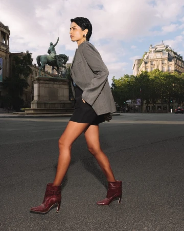 Kirandeep Chahal walks across an urban street wearing a grey blazer, black shorts, and burgundy leather ankle boots for Malone Souliers.