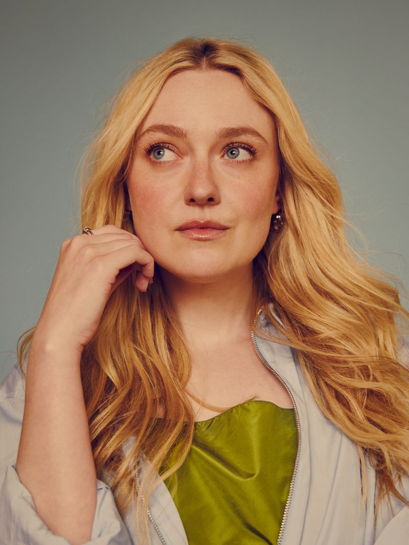 Dakota Fanning with blonde hair, wearing a light blue jacket and green top, looking away against a soft blue background.