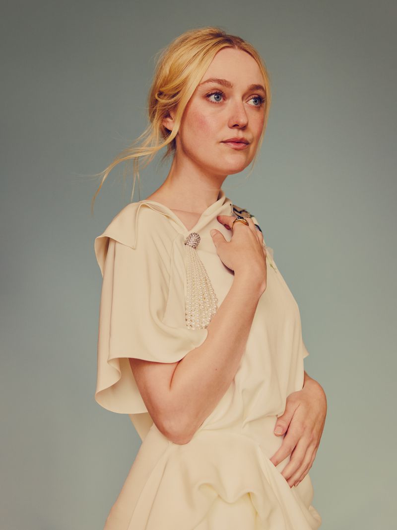Dakota Fanning with blonde hair in an updo wears a cream top with pearl fringe detail, captured in a portrait against a soft grey background.