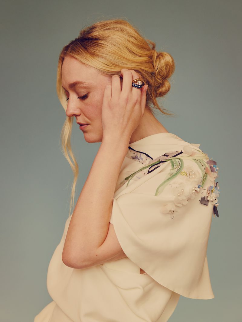 Dakota Fanning in profile view, wearing an off-white blouse with floral embroidery, touching her hair against a muted backdrop.