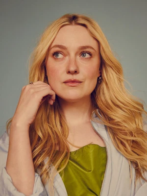 Dakota Fanning with long blonde hair wearing a green top and light jacket against a muted studio background.