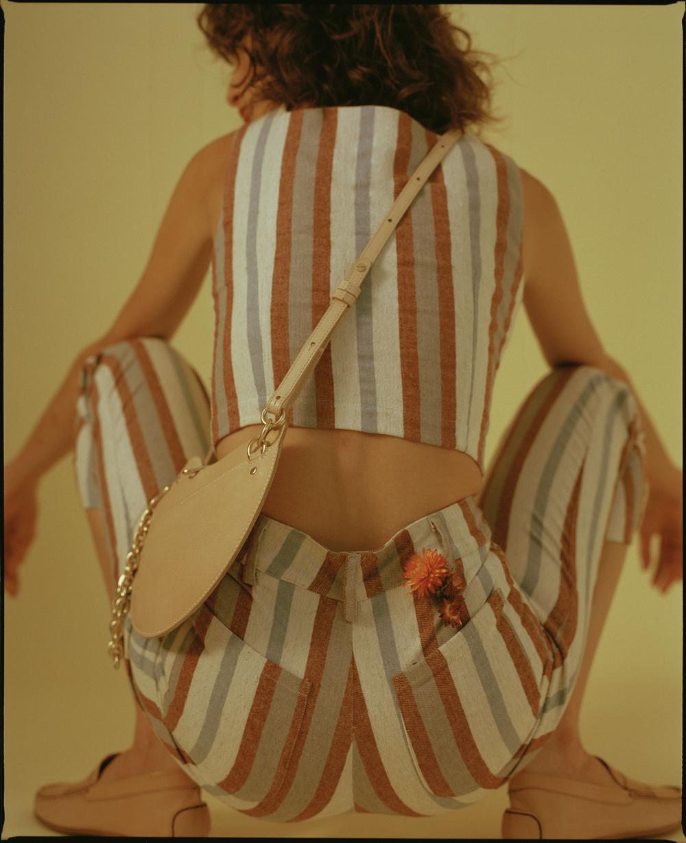 Back view of a young woman crouching, wearing striped crop top and pants, tan loafers, and carrying a matching handbag with chain.