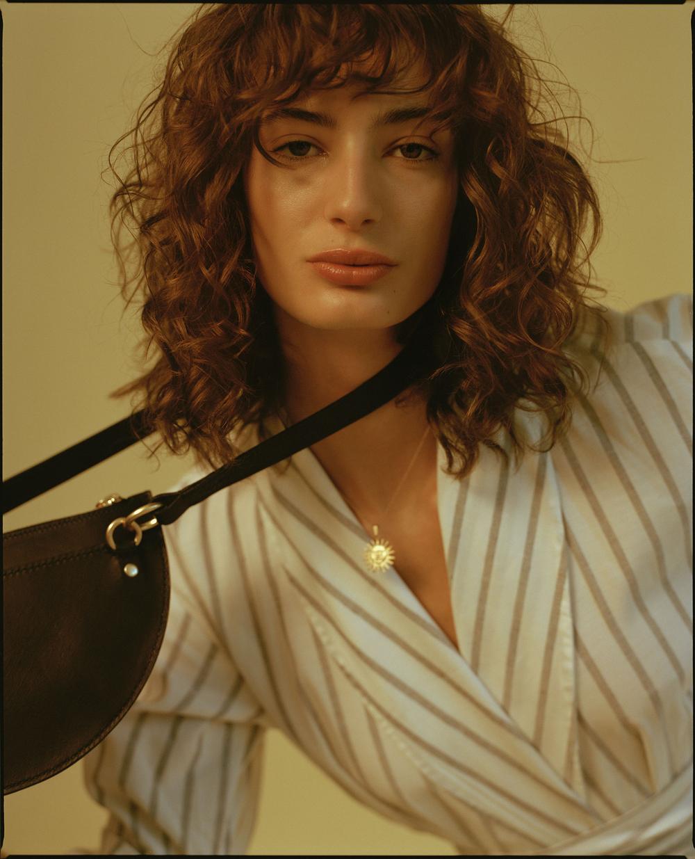 Close-up of a brunette adult woman wearing a striped shirt and a black shoulder bag, posing in front of a soft yellow background.