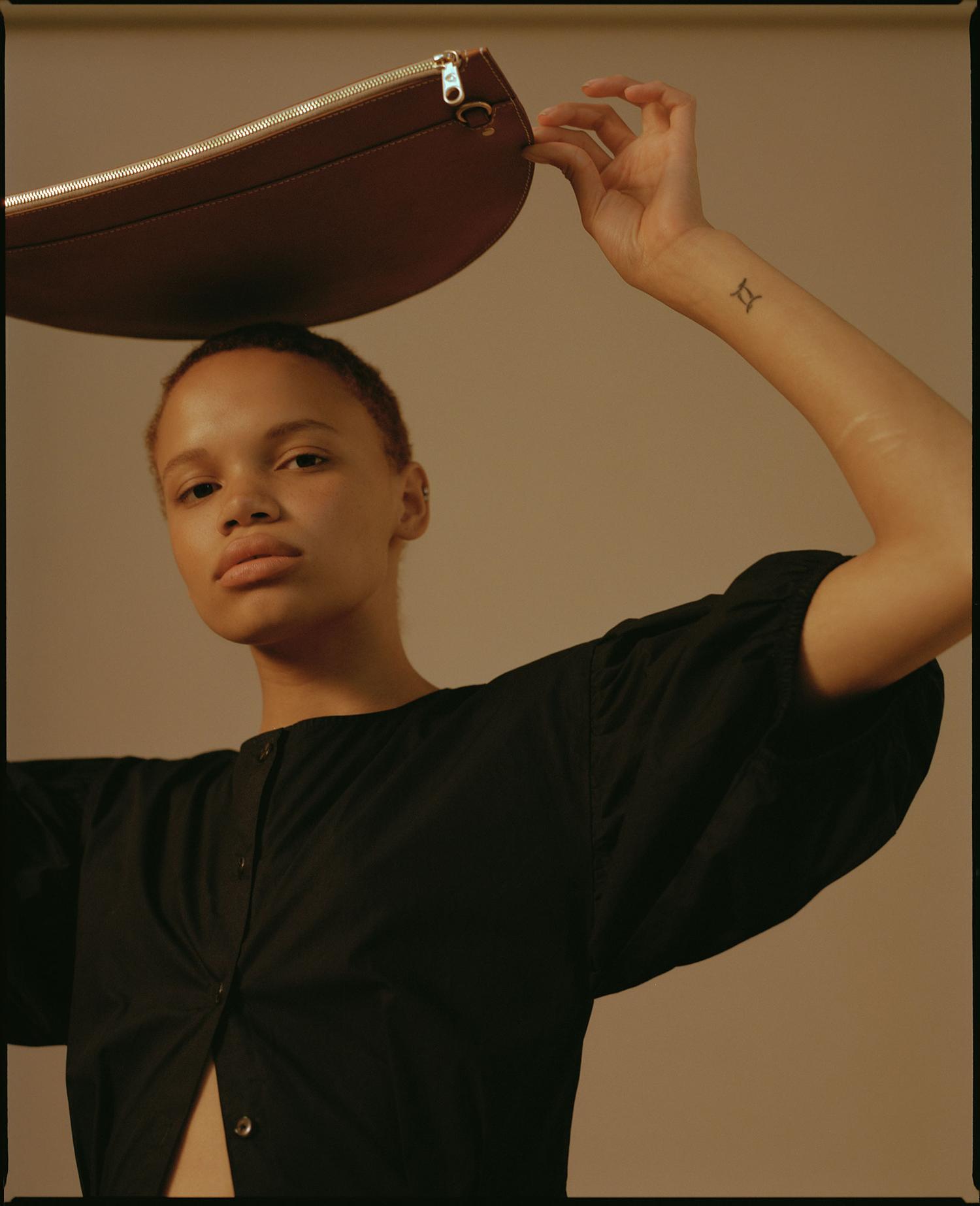 Adult woman poses in a studio with a leather bag held over her head in a fashion-forward portrait with a neutral background.