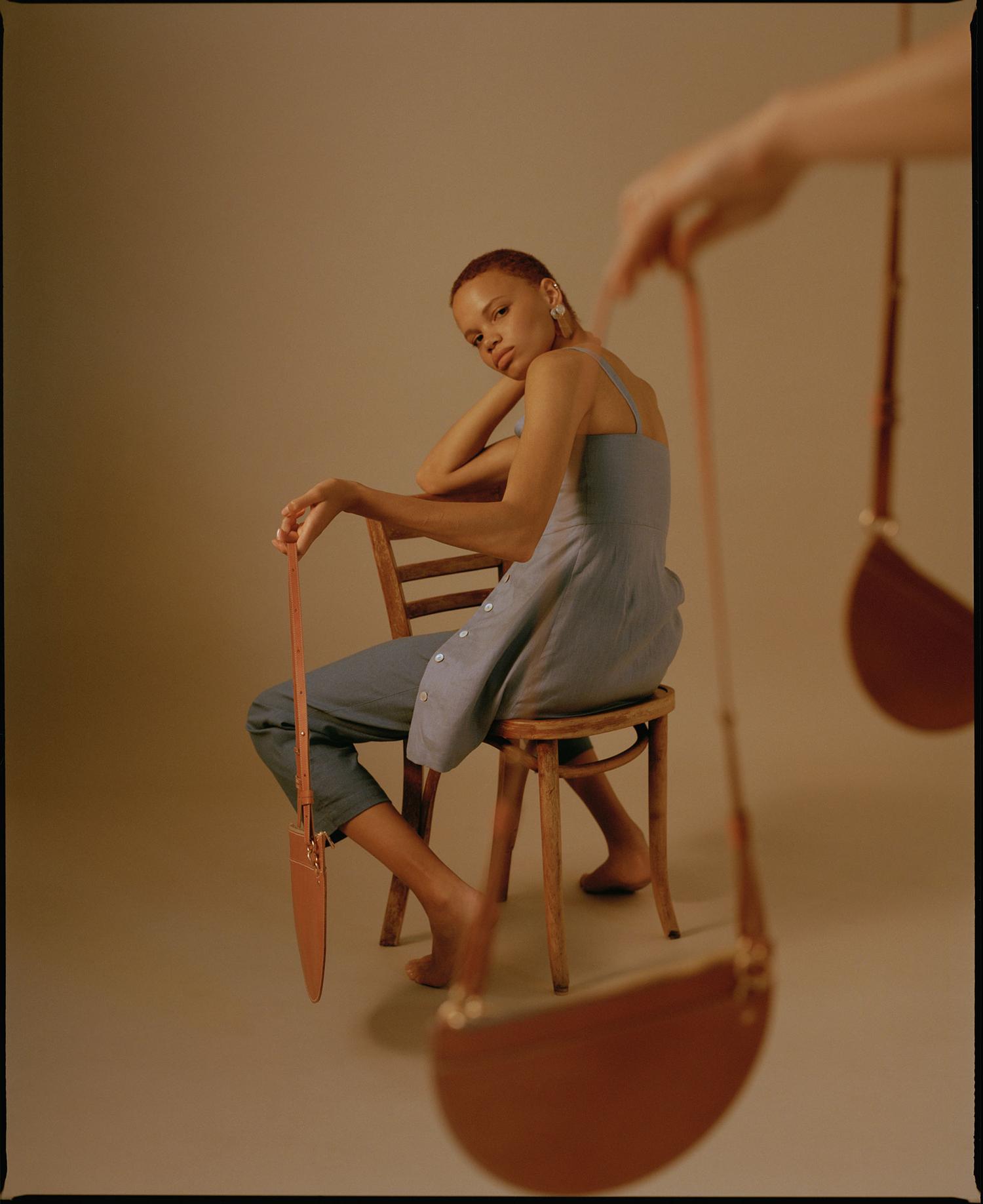 Woman in blue dress poses with leather half-moon purse on chair in studio with a second purse in the background; fashion, style.