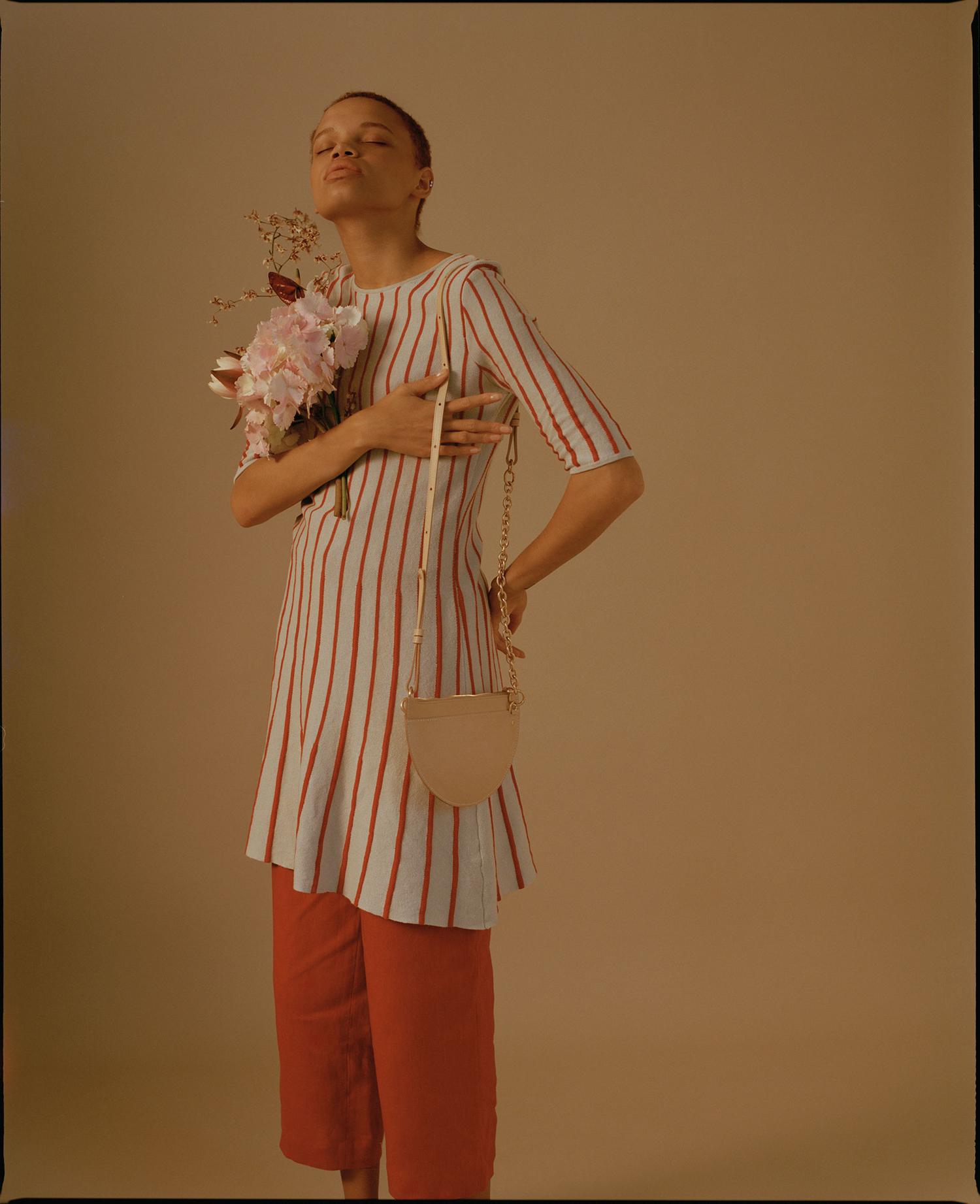 A fashion portrait of an adult woman in a striped dress and orange pants, holding flowers and a purse in a studio setting.