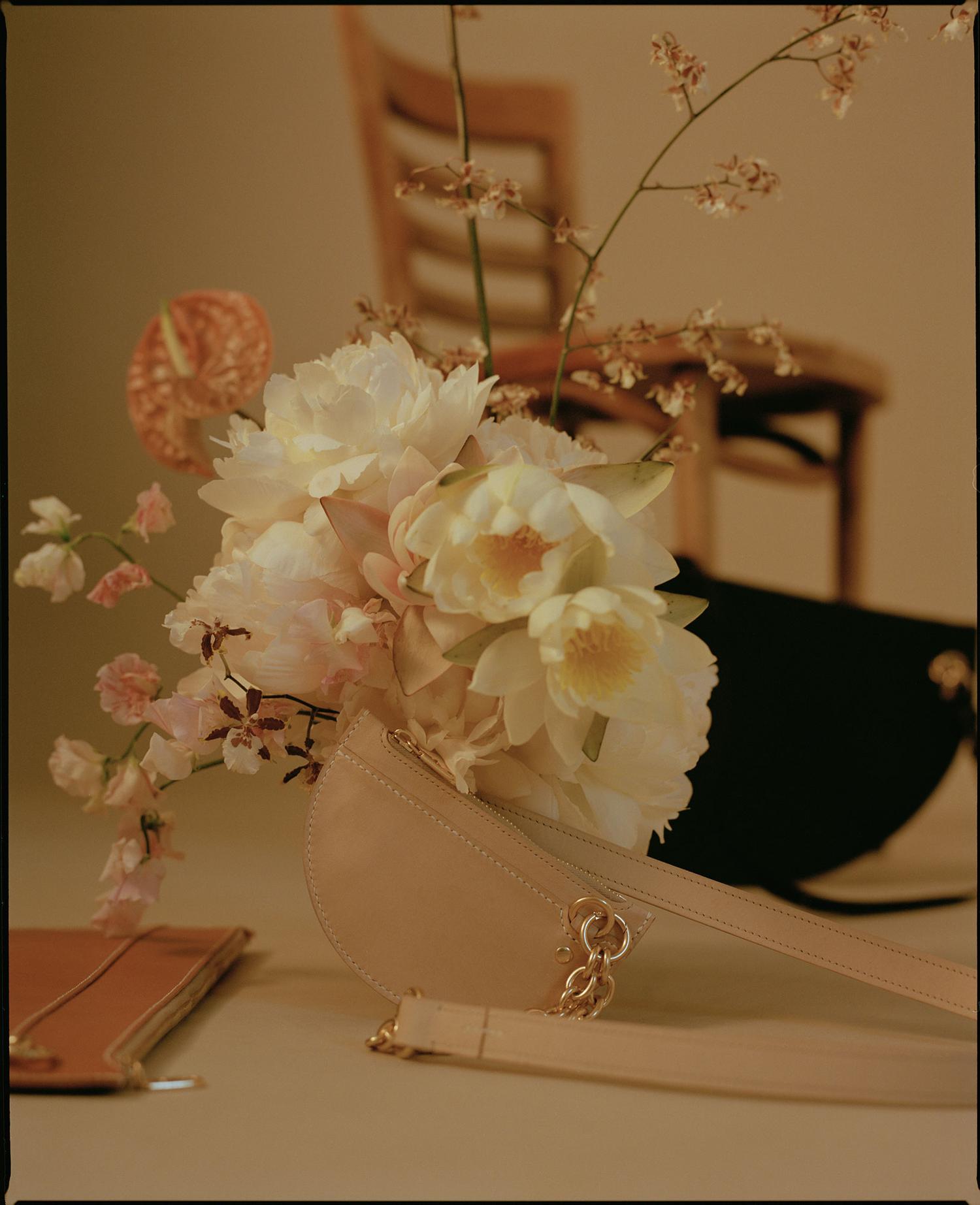 Cream-colored handbag and wallet displayed with floral arrangement in a studio setting, featuring a wooden chair in the background.