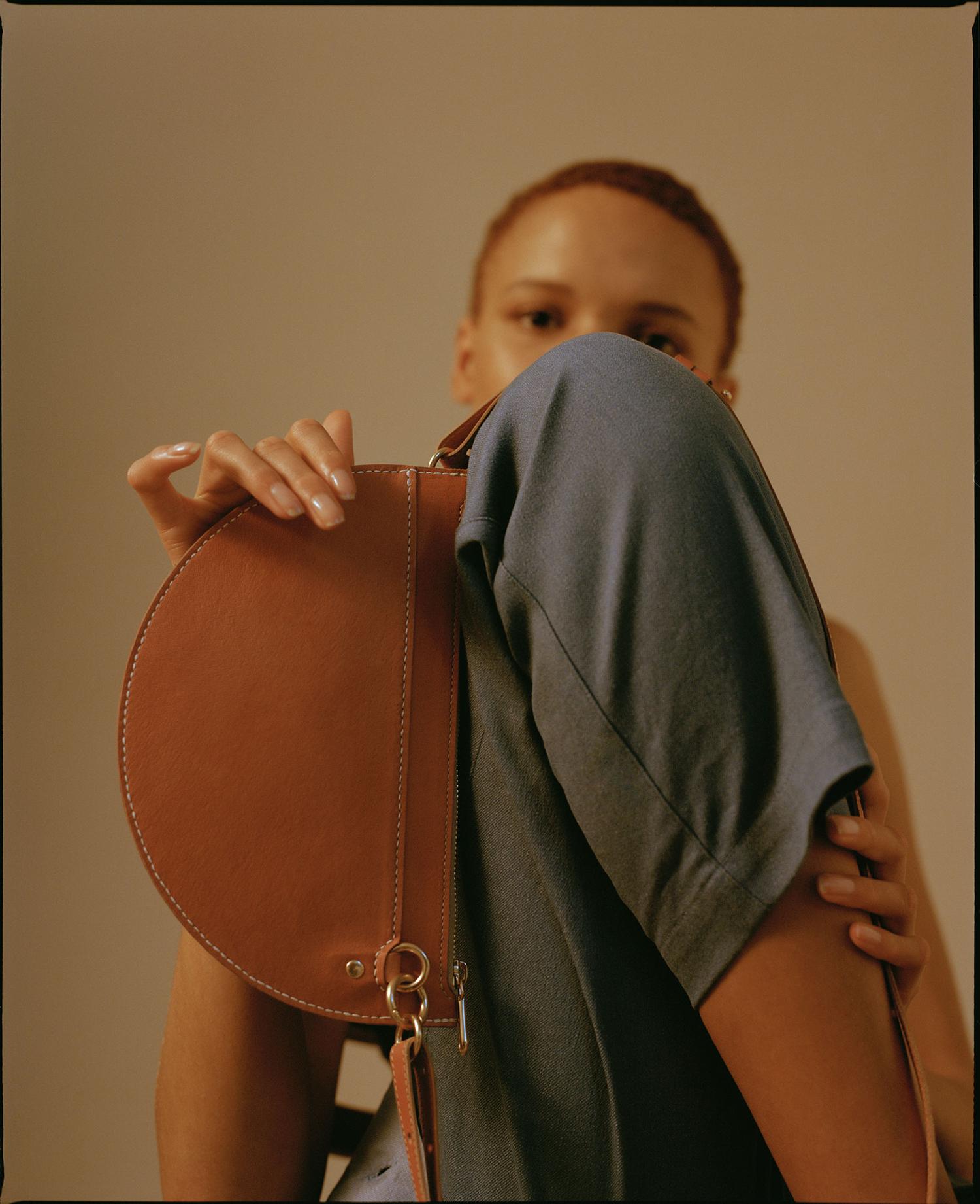 Portrait of a person with short red hair, wearing blue clothing, holding a brown leather handbag in a low-angle, indoor shot.