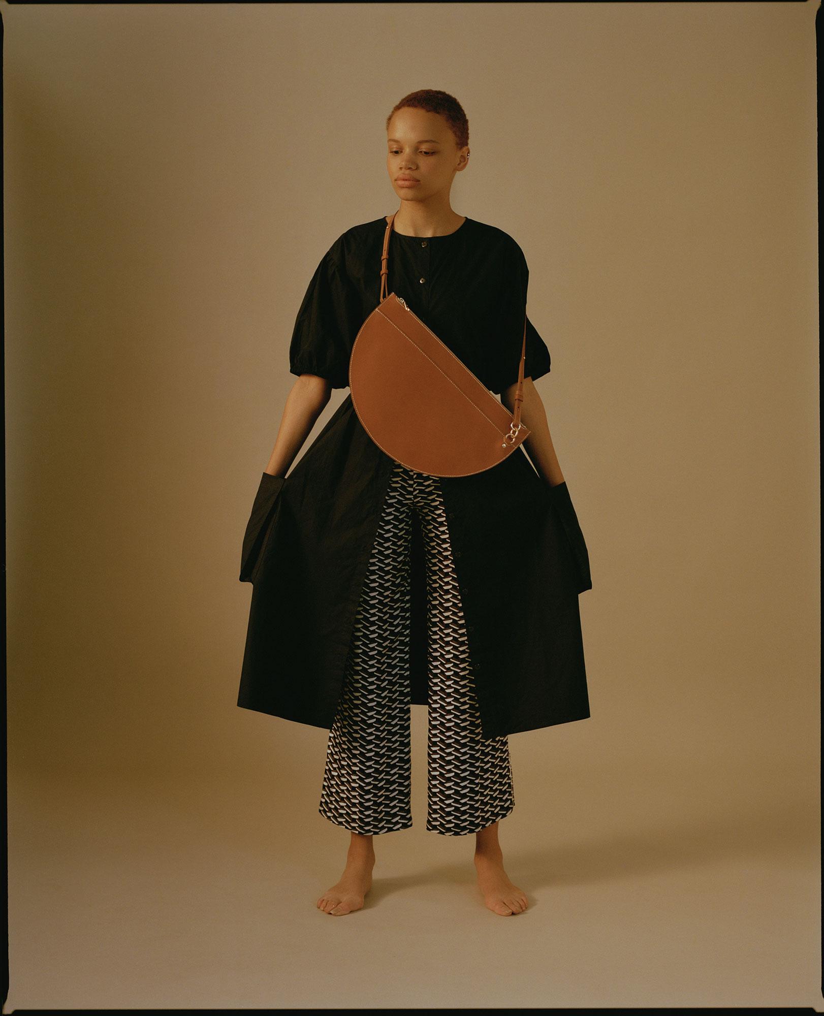 Full-shot of a slender adult female model in a black dress with a brown cross-body bag and patterned wide-leg pants, posing indoors.