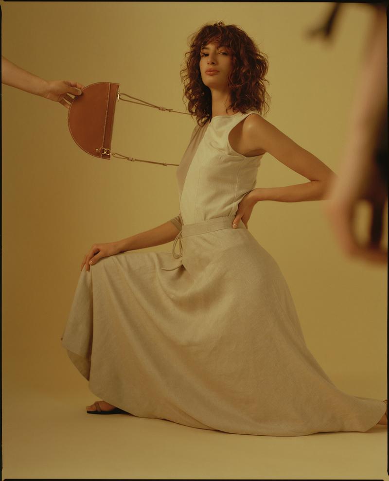 A tan handbag is presented to a woman in a beige dress. The woman is posing in front of a tan backdrop inside a photo studio.