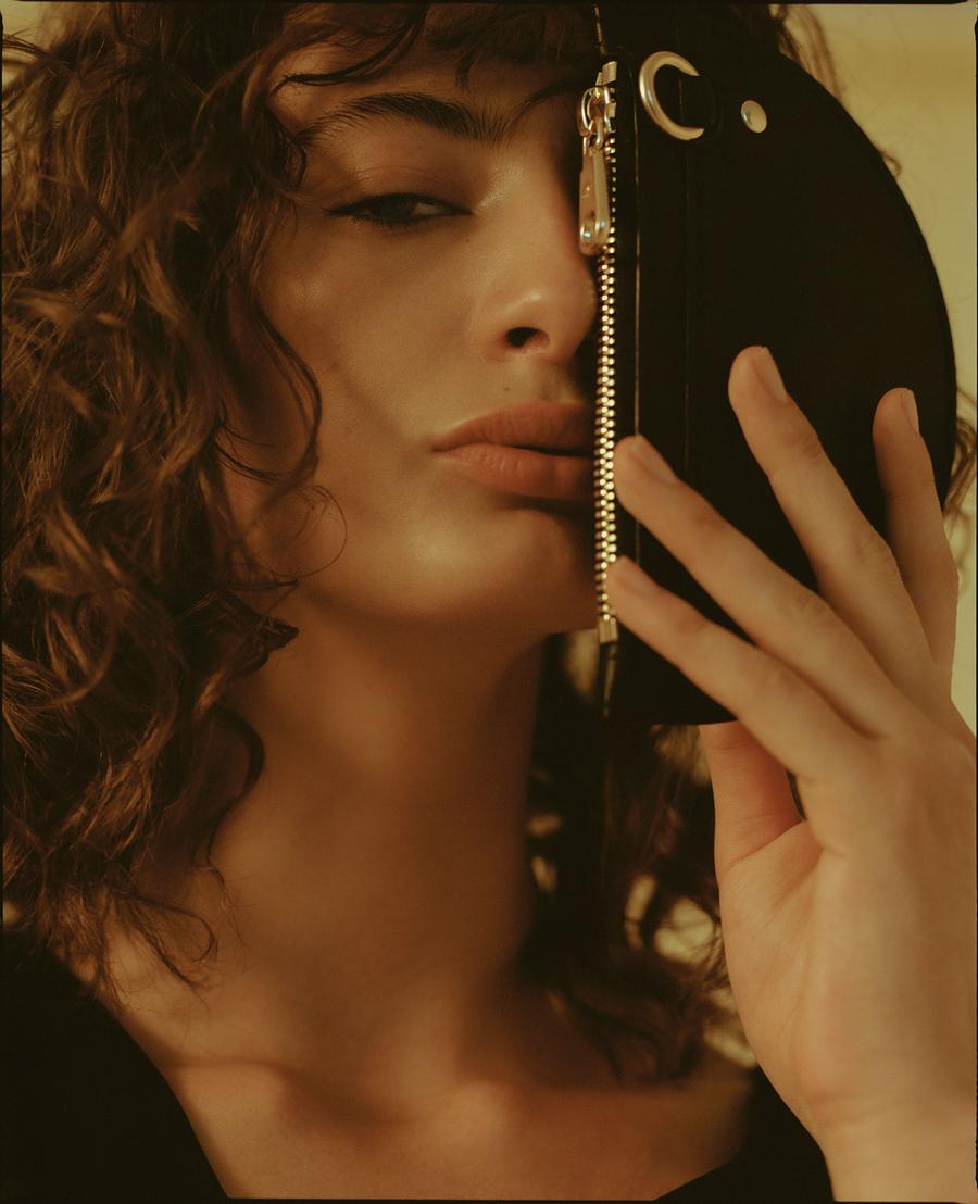 Close-up portrait of a woman with curly brown hair holding a black leather handbag half covering her face for Simétrie project.