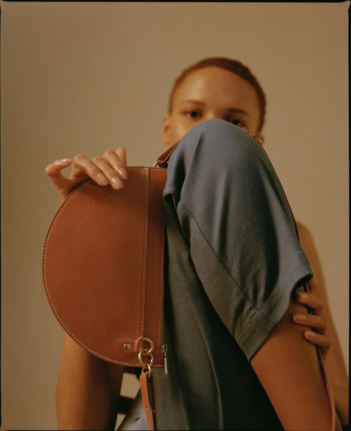 Model holding a structured brown leather saddle bag in front of her face, wearing a slate grey top against a warm neutral backdrop