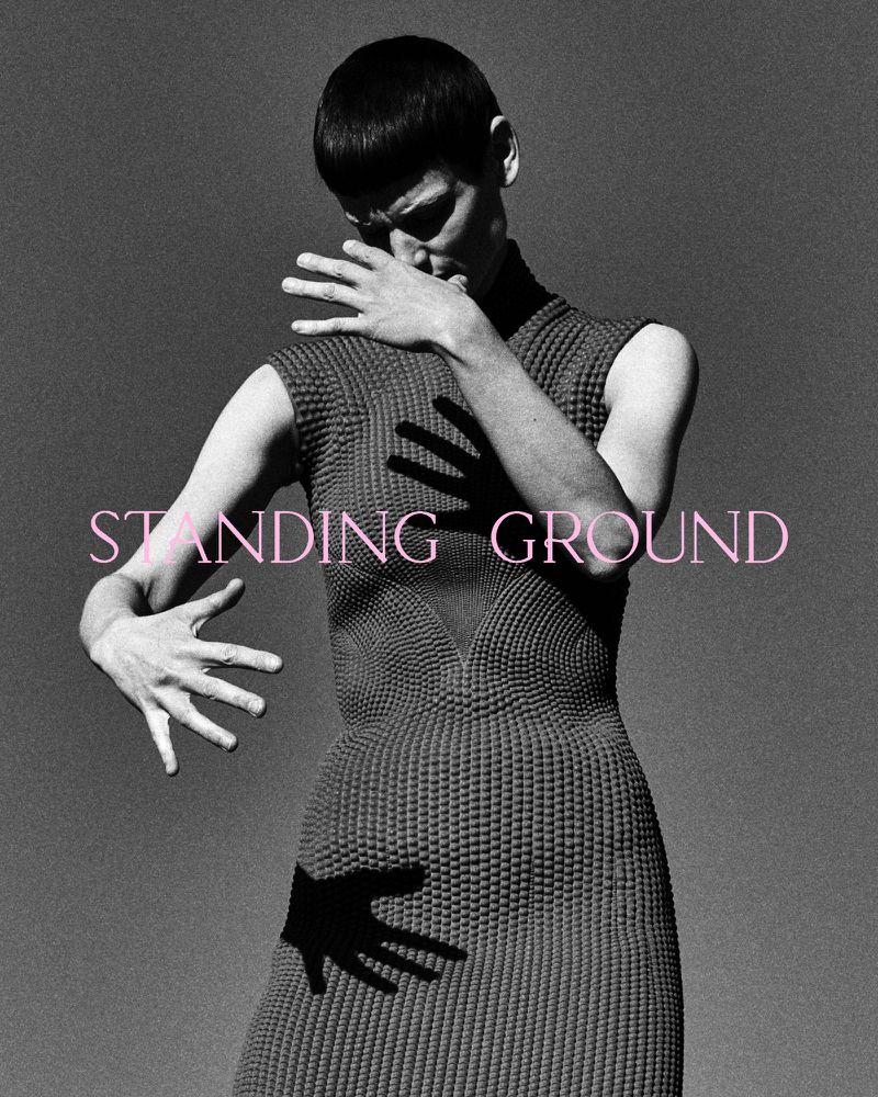 Monochrome fashion shot of androgynous model in textured grey dress posing with outstretched hands casting shadows on dress and backdrop.