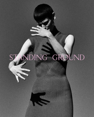 Monochrome fashion shot of androgynous model in textured grey dress posing with outstretched hands casting shadows on dress and backdrop.