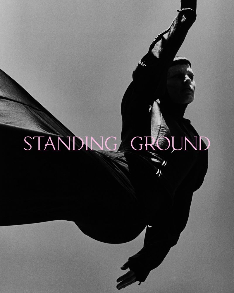 Dramatic black-and-white fashion shot: a person in motion against a bright sky, wearing dark, flowing clothing with graphic text overlay.