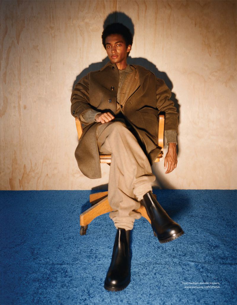 Model Stan Taylor seated in a wooden office chair wearing an olive coat, beige trousers, and black leather boots against plywood.