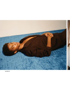 Model Stan Taylor wearing an open brown suit lying down on a bright blue shag carpet against a plain wall.