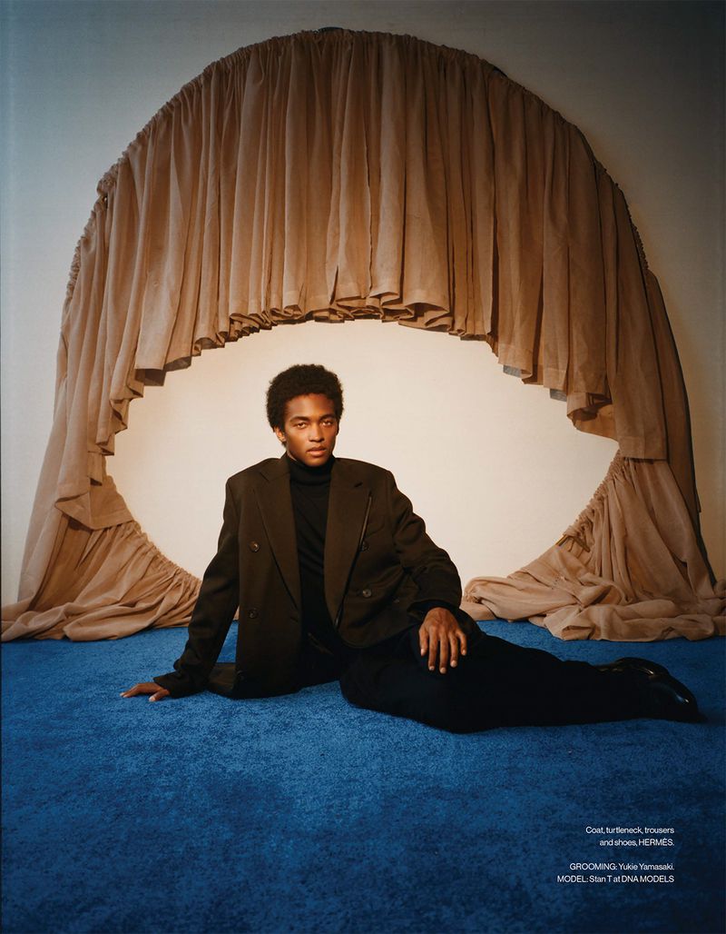 Model Stan Taylor reclining on blue carpet wearing a dark blazer and black turtleneck in front of a draped beige arch backdrop.