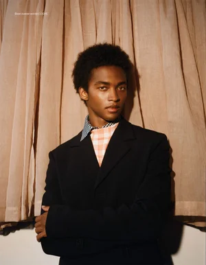 Model Stan Taylor in a black blazer layered with a checkered knit top against draped beige fabric curtains.