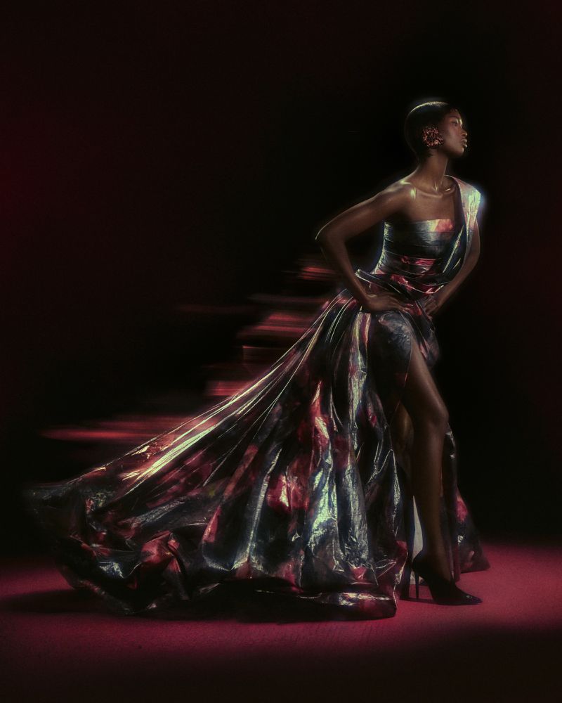 Model Nyarach Abouch Ayuel poses in an iridescent metallic high-slit evening gown against a dark background with red ambient light.