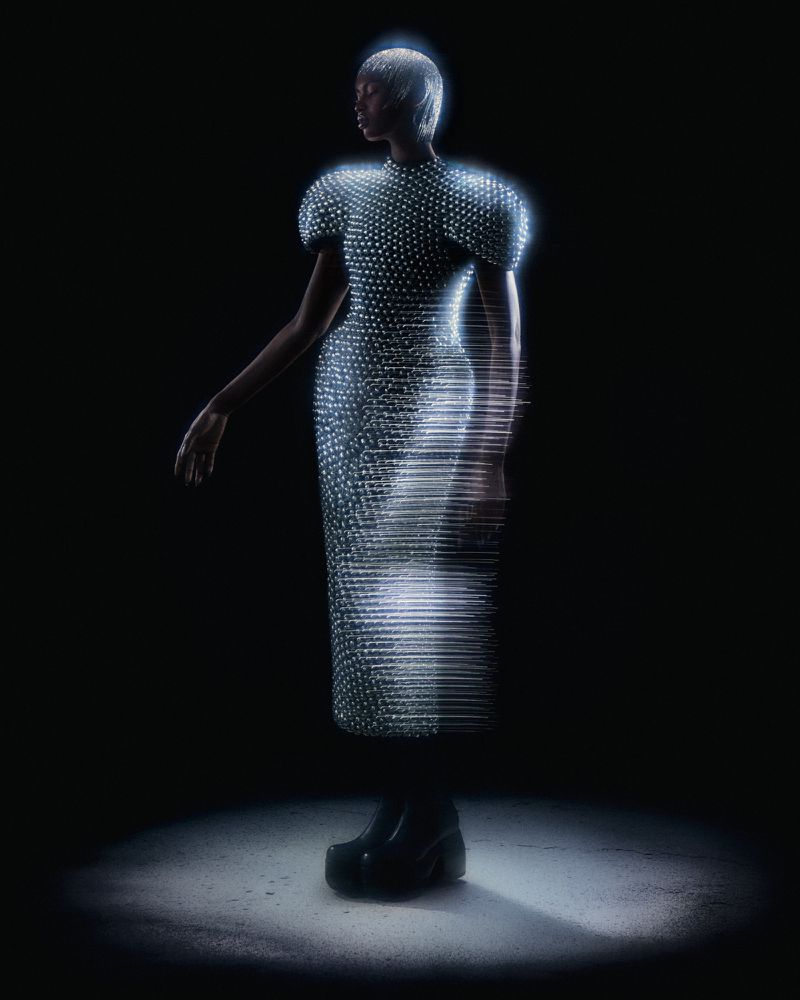 Model Nyarach Abouch Ayuel standing in a spotlight wearing an illuminated silver dress with digital scan lines on a dark background.