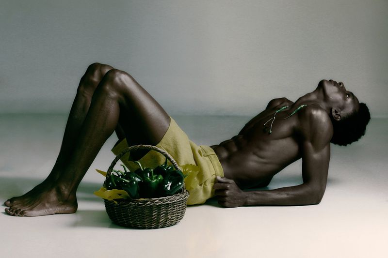 Shirtless Black man in yellow shorts reclining on the floor beside a basket filled with green peppers in a studio setting.