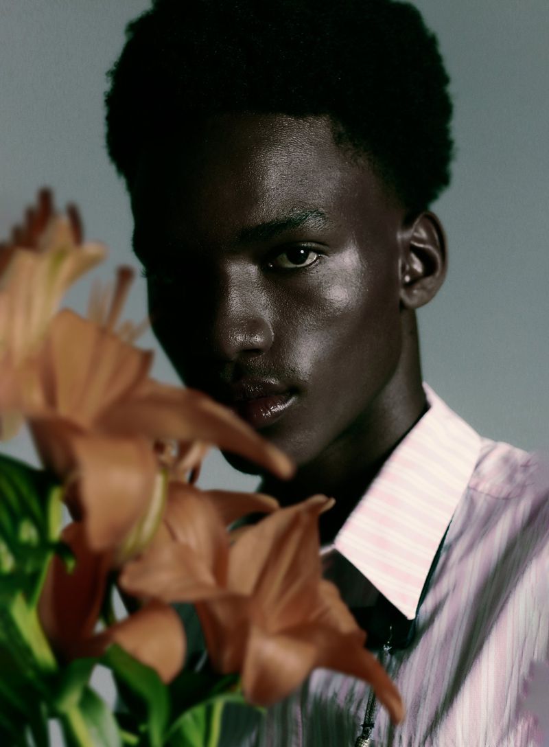 Close-up portrait of a Black man in a pink striped shirt framed by brown lily flowers against a grey background.