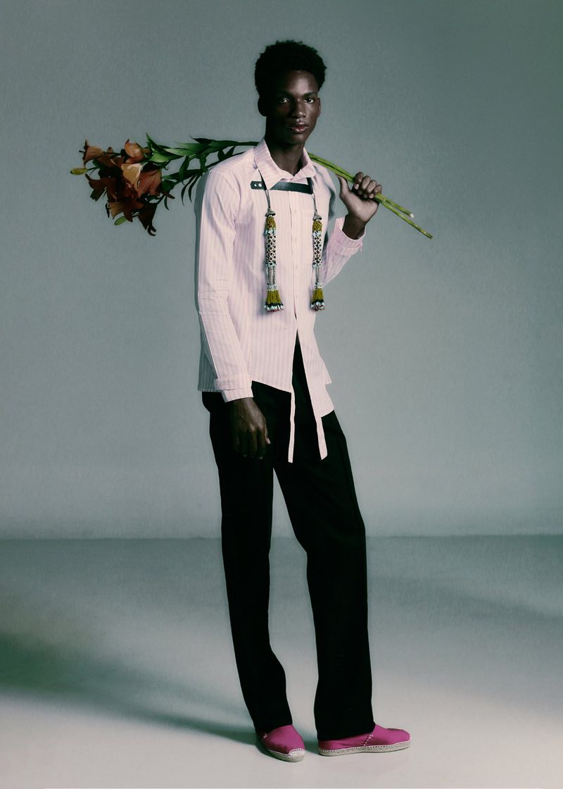 A young Black man in a pale pink shirt and black trousers stands holding a long stem of brown lilies over his shoulder in a studio setting.