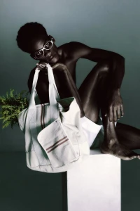 Black model wearing sunglasses and white shorts seated on a white pedestal holding a canvas tote bag with greenery in a studio setting.