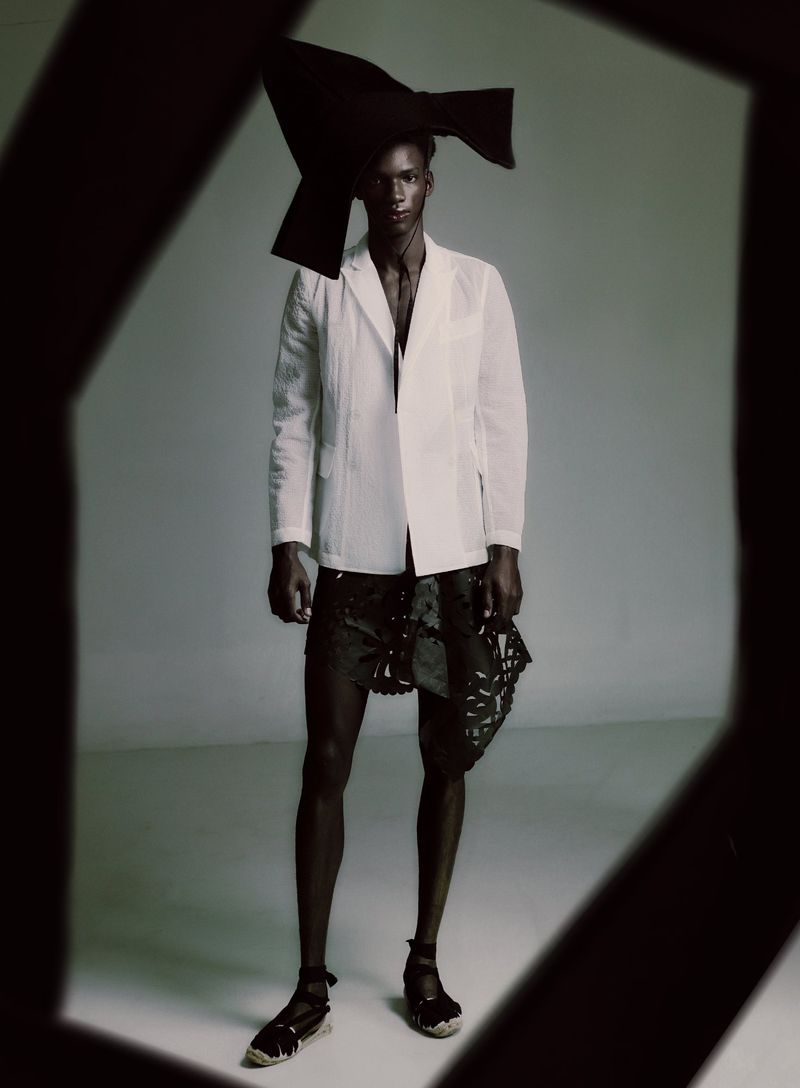 Black male model in a white blazer, black headpiece, and espadrilles standing in a studio framed by dark geometric foreground.
