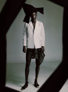 Black male model in a white blazer, black headpiece, and espadrilles standing in a studio framed by dark geometric foreground.