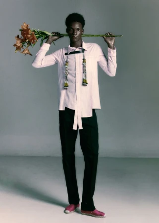 A Black male model in a pale pink shirt, black trousers, and pink espadrilles holding an orange lily stem behind his neck in studio.
