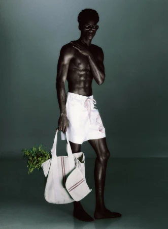 Shirtless Black man wearing white shorts and dark glasses holding a canvas tote bag filled with green foliage against a dark backdrop.