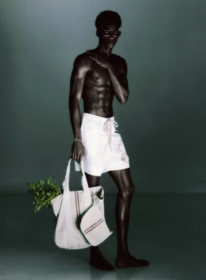 Shirtless Black man wearing white shorts and dark glasses holding a canvas tote bag filled with green foliage against a dark backdrop.