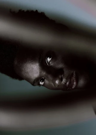 Low-key close-up portrait of a Black model looking into the lens, framed tightly by blurred foreground shapes.