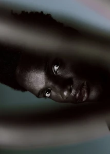 Low-key close-up portrait of a Black model looking into the lens, framed tightly by blurred foreground shapes.