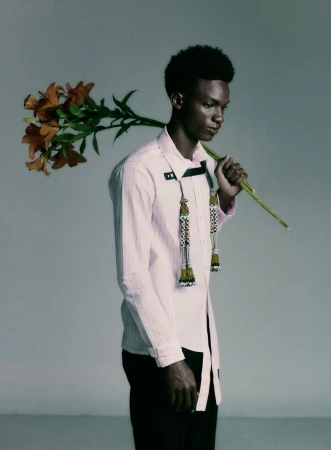 A young Black man in a pale pink shirt holds a stem of orange lilies over his shoulder against a plain grey studio backdrop.