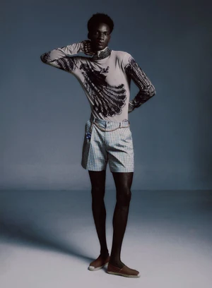 Black male model in a printed long-sleeve shirt and check shorts standing against a grey studio backdrop.
