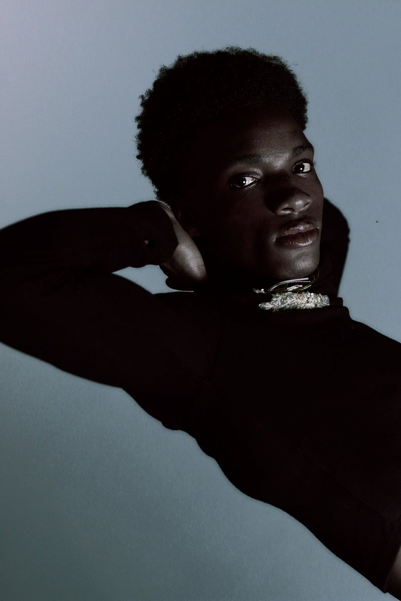 Black man posing with arms behind head wearing a black top and textured silver choker against a cool grey background.