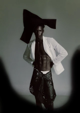 Black male model in an open white blazer and sculptural black headpiece against a grey background with vignetted edges.