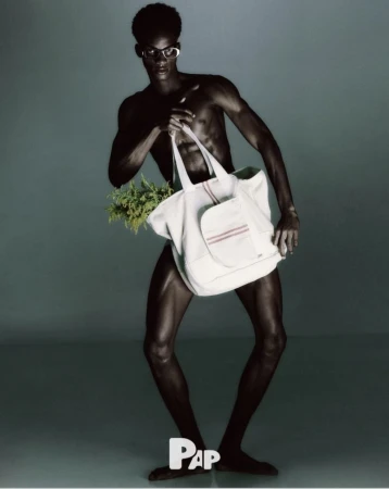 Black male model wearing sunglasses and posing while holding a large white canvas tote bag filled with green foliage in a studio setting.