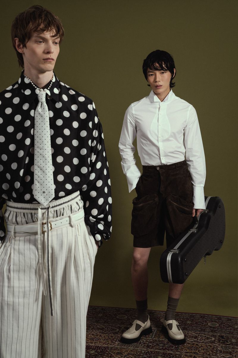 Two male models posing in studio, one in a polka dot shirt and pinstripe pants, the other in a white shirt and shorts holding a guitar case.