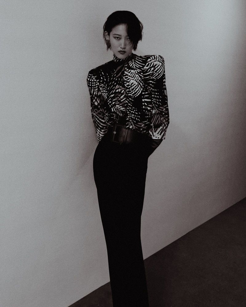 Monochrome studio shot of a stylish adult female model wearing a patterned high-neck blouse, wide belt, and long black skirt, posing elegantly.