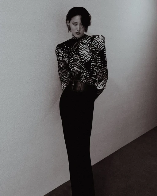 Monochrome studio shot of a stylish adult female model wearing a patterned high-neck blouse, wide belt, and long black skirt, posing elegantly.