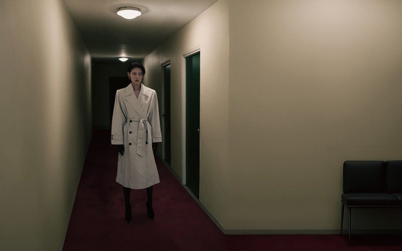 Woman in a belted trench coat stands in a long hallway with dark doors and dim lighting, creating a mysterious, dramatic scene.