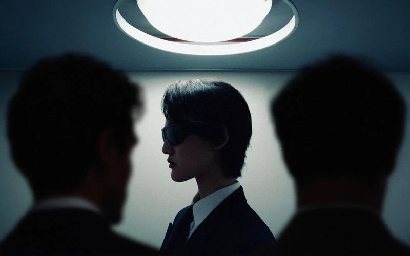 An androgynous person in a suit, wearing sunglasses, is framed by two blurred silhouettes, highlighted by overhead lighting.