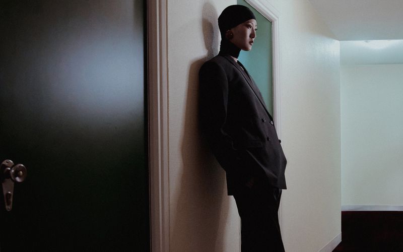 Woman in a black suit leans against a wall in a hallway, with a dark door on the left and a light-colored wall with a doorway on the right.