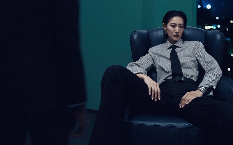 A woman with short black hair is sitting in a black leather chair wearing a pinstripe shirt, tie, and dark pants in an indoor setting.