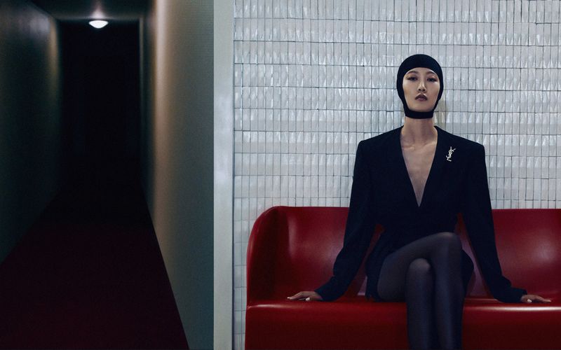 A slender woman in a black suit and head covering sits on a red bench against a white tiled wall, with a dark corridor visible.