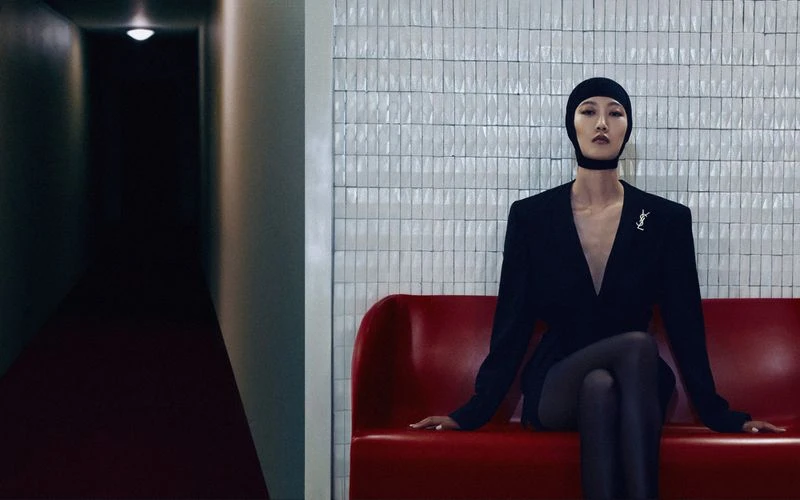 A slender woman in a black suit and head covering sits on a red bench against a white tiled wall, with a dark corridor visible.